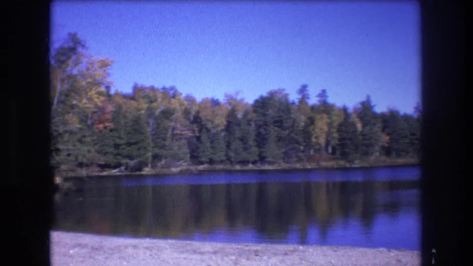1975 Baxter, Maine 8mm frame showing a wider lakeside view with calm reflective water, mixed evergreen and autumn foliage, blue sky, and sandy foreground.
