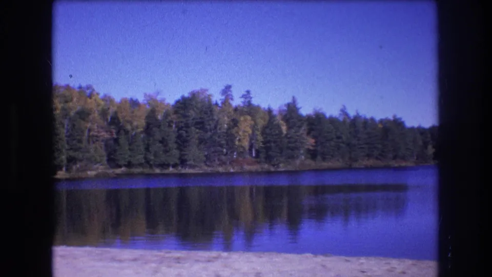 1975 Baxter, Maine 8mm frame of a calm blue lake bordered by dense forest, with tree reflections, clear sky, sandy foreground, and dark film edges.