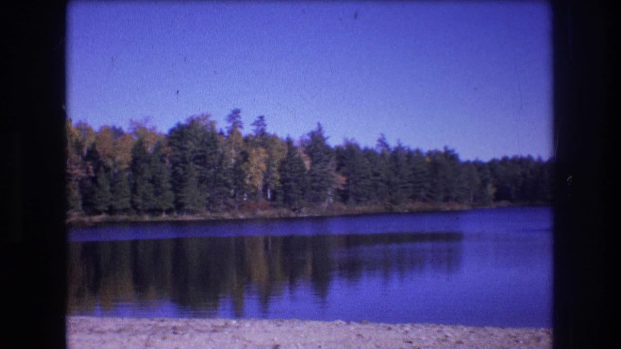 1975 8mm home-movie frame from Baxter, Maine, showing calm lake water, a sandy beach edge, forested shoreline, tree reflections, and a clear blue sky.
