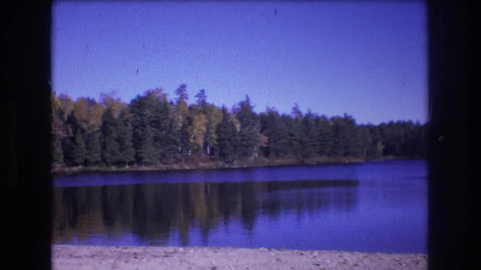 1975 8mm home-movie frame from Baxter, Maine, showing calm lake water, a sandy beach edge, forested shoreline, tree reflections, and a clear blue sky.