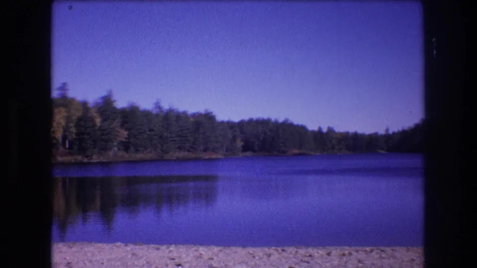 1975 Baxter, Maine 8mm frame showing a wider stretch of blue lake, distant forested banks, soft reflections, and a sandy or pebbled beach at the bottom of the frame.