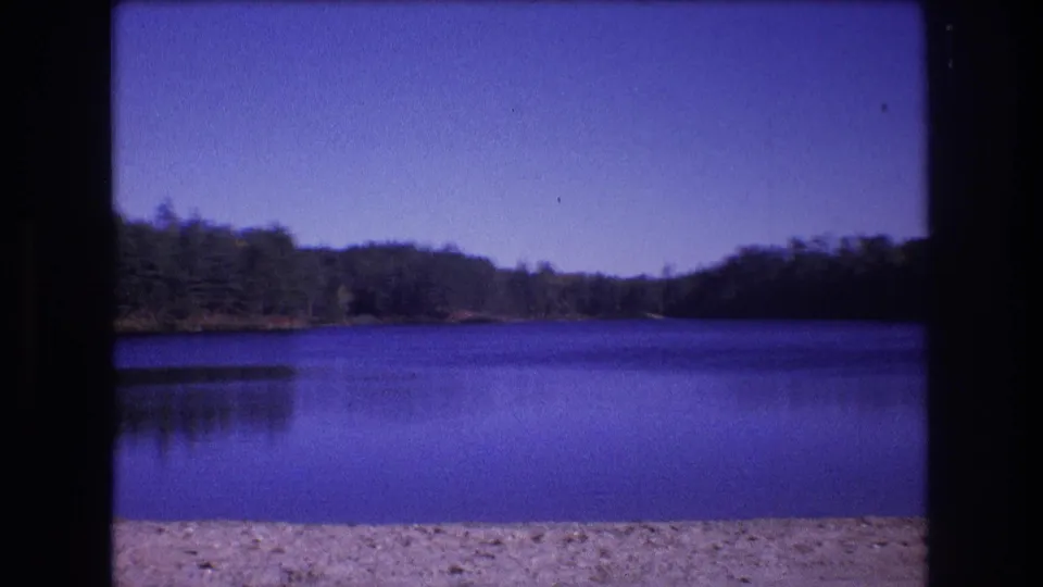 Final 1975 8mm frame from Baxter, Maine, looking across open blue lake water toward a dark forested shoreline with a sandy foreground and clear sky.