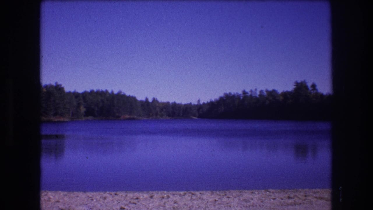 1975 Baxter, Maine 8mm frame showing a calm lake from a sandy beach, with a dark wooded shoreline under a clear sky and a pronounced purple-blue film cast.