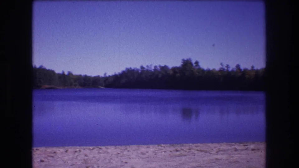 1975 Baxter, Maine home-movie frame with still lake water, sandy shoreline in front, dense forest across the water, and visible 8mm scan borders.