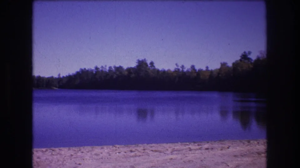 1975 Baxter, Maine 8mm frame showing the same lakeside scene from a fixed position, with beach texture in the foreground and forest reflected in the calm water.