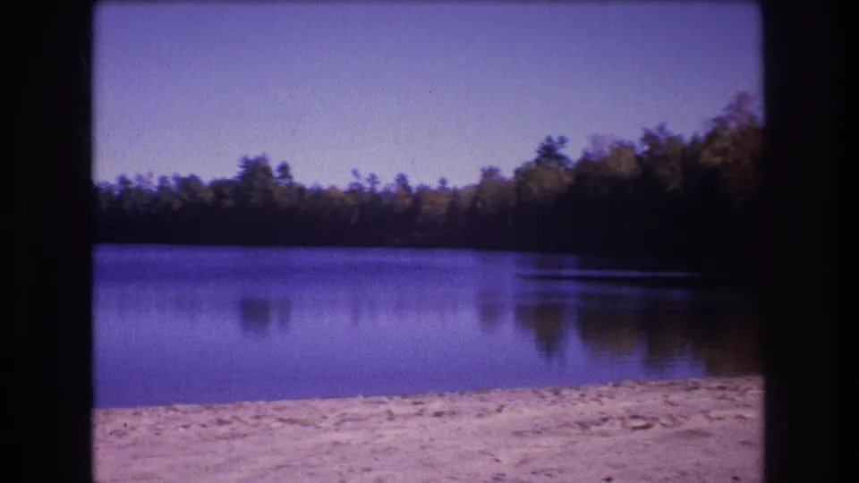 1975 Baxter, Maine frame of a quiet lake and tree-covered shore, with the sandy beach in front and darker reflected trees along the right side of the water.