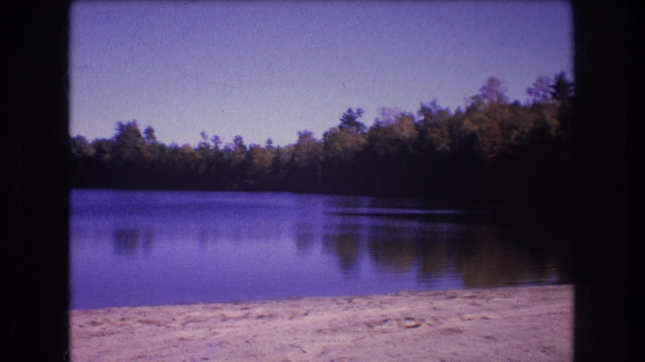 1975 Baxter, Maine 8mm frame showing a calm wooded lake with a pale sandy shoreline and clear sky.