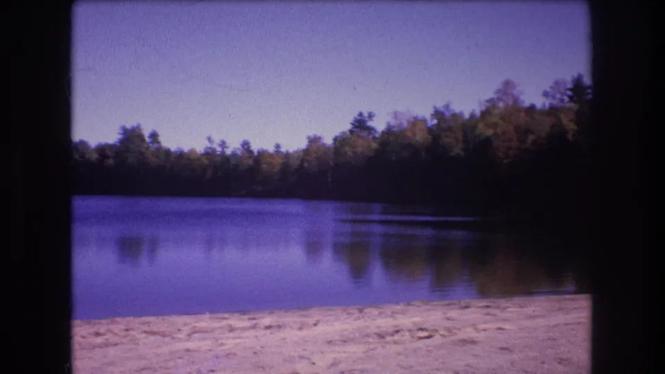 1975 Baxter, Maine 8mm frame showing a calm wooded lake with a pale sandy shoreline and clear sky.