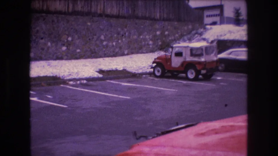 1975 Baxter, Maine 8mm frame showing a red Jeep-style vehicle and another car parked below a stone wall near a light-colored building.