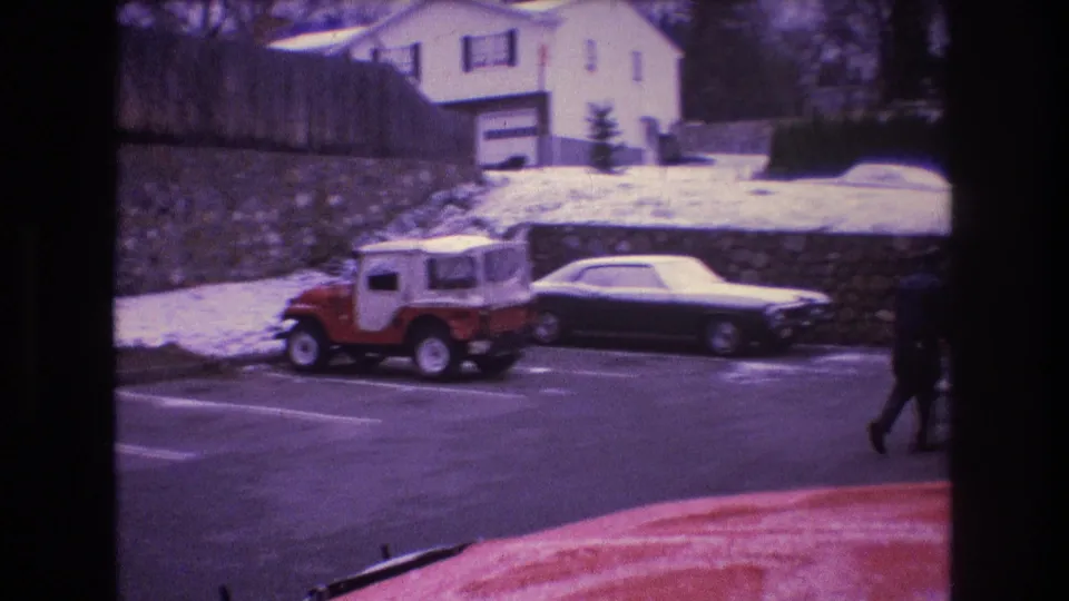 1975 Baxter, Maine frame showing parked vehicles, a stone retaining wall, a light-colored house or garage, and a partly visible person at the right edge.