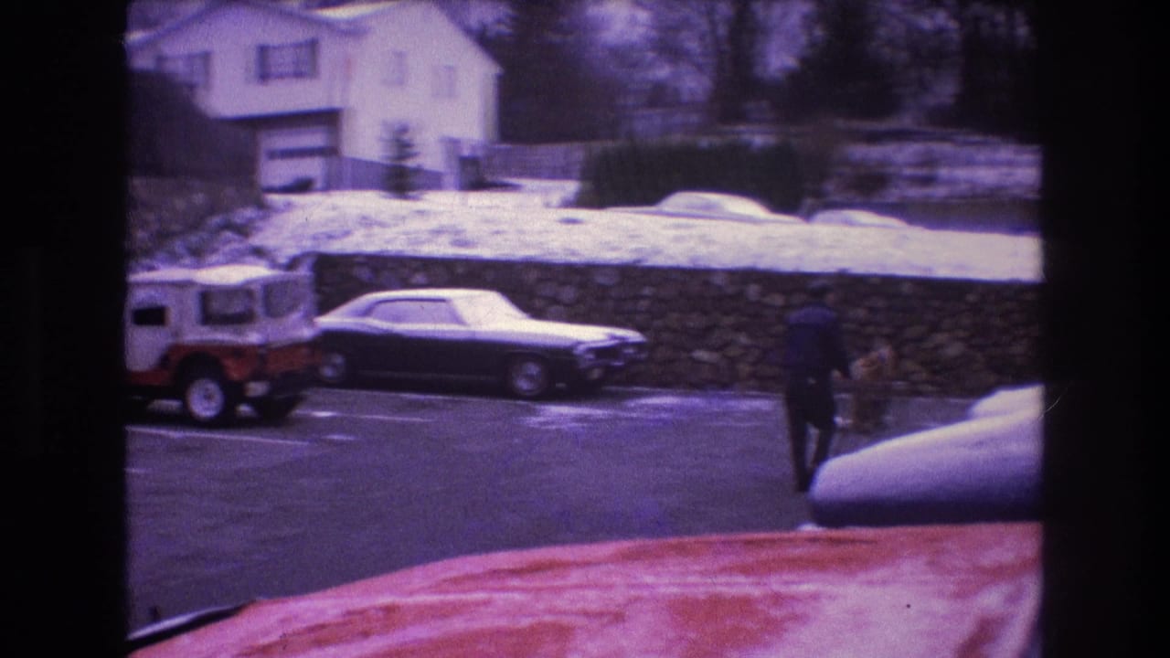 In Baxter, Maine in 1975, a distant man in dark winter clothing walks through a snowy parking lot with parked cars, a stone wall, and houses beyond.