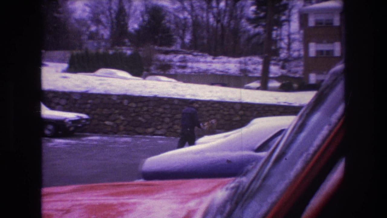 Distant man walking near snowbanks with parked cars and houses behind him.