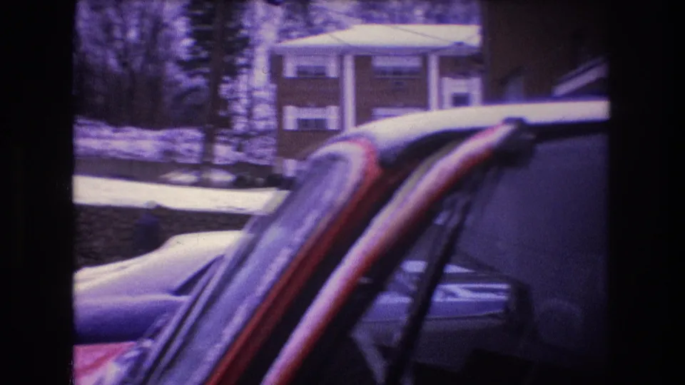 The camera frames the snow-dusted windshield area of a red car in a Baxter, Maine parking lot, with another car and residential buildings beyond.