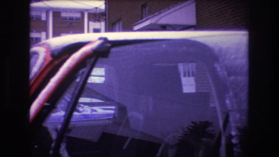 A close handheld view shows a red vehicle windshield and roof dusted with snow in the Baxter, Maine winter parking area, with a residential building and another car visible behind it.