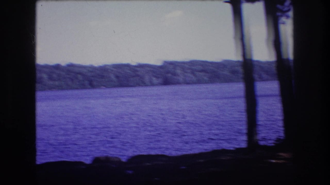 1975 8mm home-movie frame from Baxter, Maine, showing rippled lake water, a wooded far shore, shoreline rocks, and dark tree trunks in the foreground.