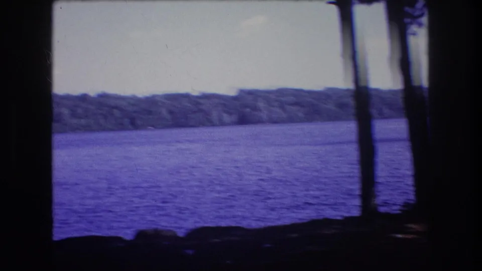 1975 8mm home-movie frame from Baxter, Maine, showing rippled lake water, a wooded far shore, shoreline rocks, and dark tree trunks in the foreground.