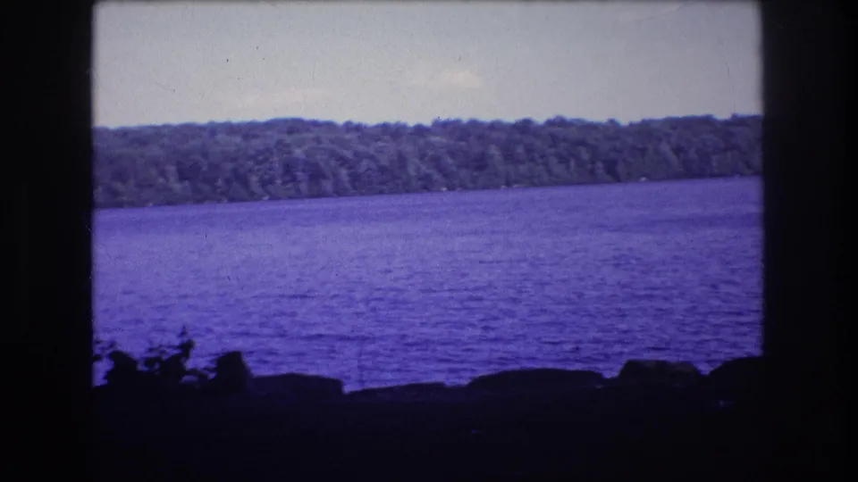 1975 8mm frame showing Baxter, Maine lake water under a pale sky, with a continuous treeline across the far shore and dark rocks in the foreground.