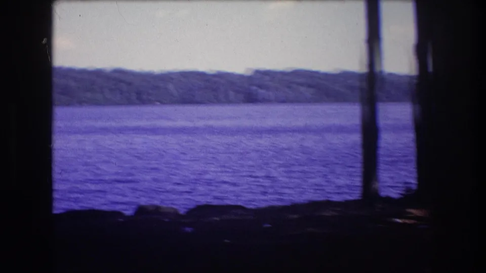 A short 8mm home-movie view from Baxter, Maine, with lake water, distant wooded shoreline, foreground tree trunks, and rocks along the lower frame.