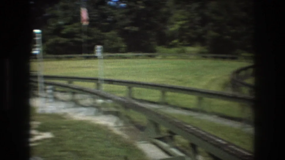 1975 8mm frame from Baxter, Maine showing a green wooden ride track curving through grass, fencing, gravel, and trees from a moving visitor viewpoint.