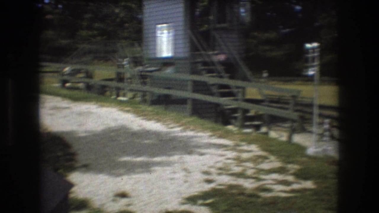 1975 8mm frame from Baxter, Maine with a small wood-sided structure, stairs, pale gravel, fencing, and a green ride track, with a partial passenger silhouette near the foreground.