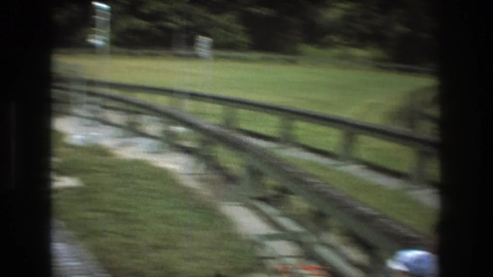 Motion-blurred 1975 8mm view in Baxter, Maine of a green wooden ride track, rail fencing, grass, gravel, and trees as the camera moves past.