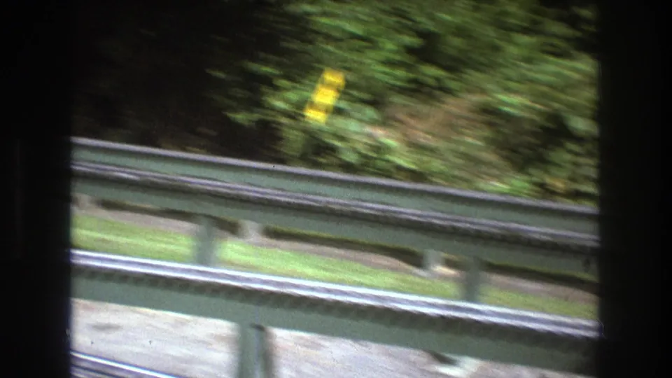 1975 8mm frame from Baxter, Maine showing a roadside guardrail or trackside rail, dense green trees, pale ground, and a blurred yellow sign during camera movement.