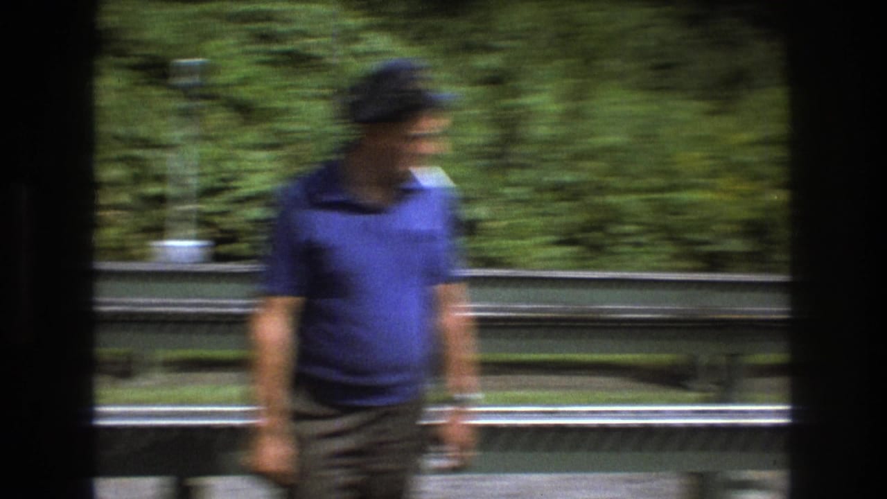 In 1975 Baxter, Maine home-movie footage, an adult man in a blue polo shirt and cap appears blurred while moving beside outdoor miniature railway tracks.