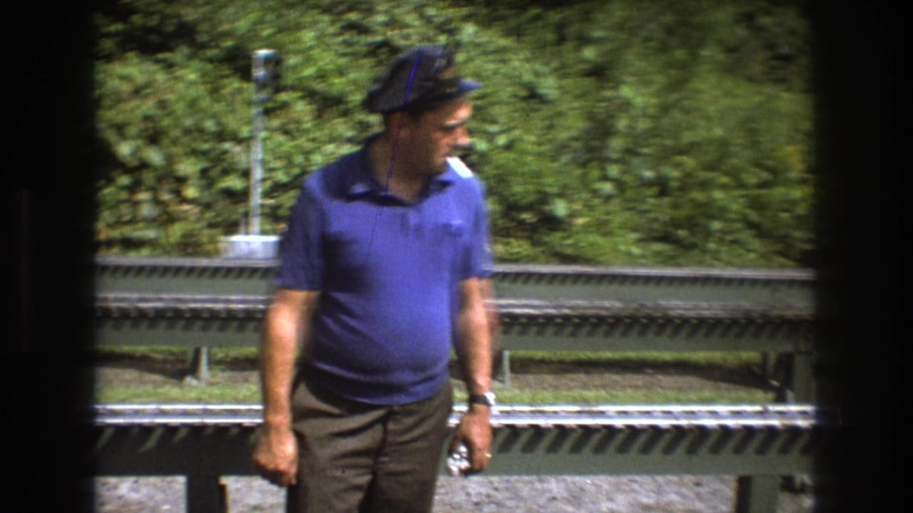 Man in a blue polo shirt and cap stands beside miniature railway tracks outdoors.
