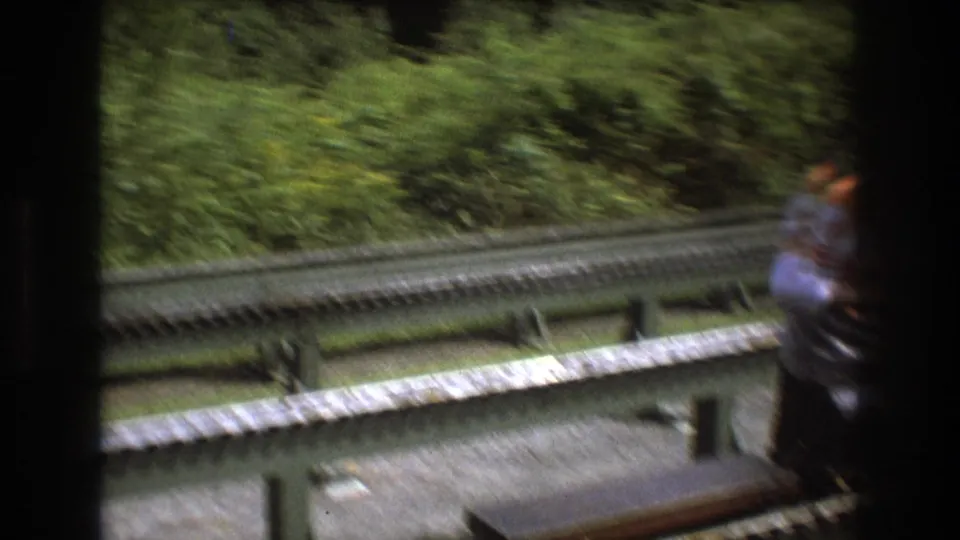 A handheld 8mm view from the Baxter, Maine miniature railway scene shows rails, guardrails, foliage, and a partially visible person moving near the lower edge of the frame.