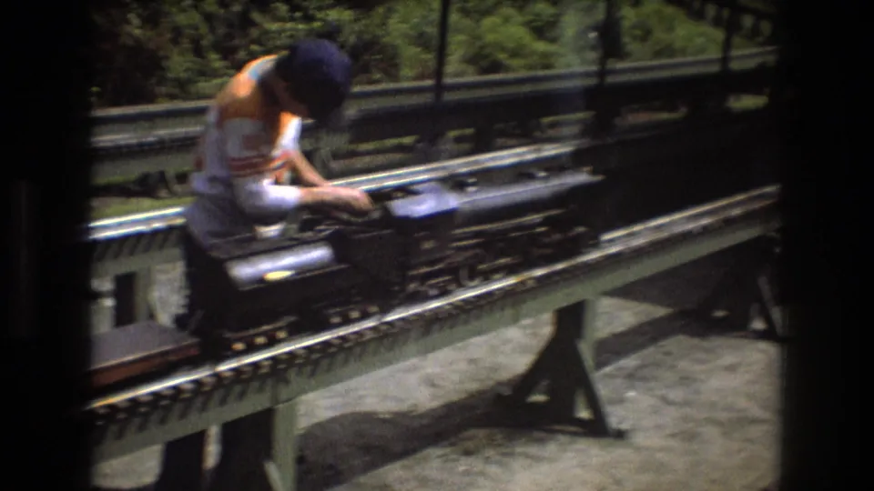 In 1975 Baxter, Maine footage, an adult man in a sleeveless striped shirt leans over a black miniature steam locomotive on raised outdoor tracks.