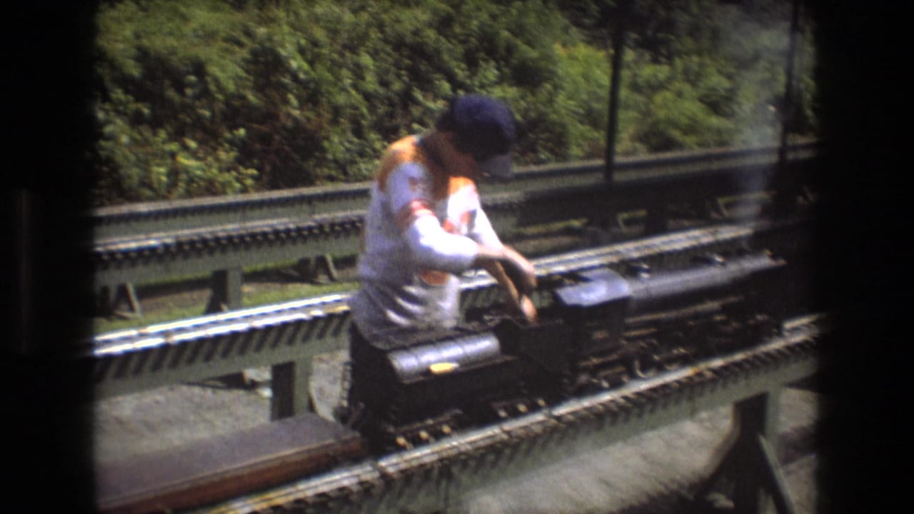 In this 1975 Baxter, Maine home-movie frame, a man in a white shirt and cap bends over a small steam locomotive on elevated miniature railway tracks.