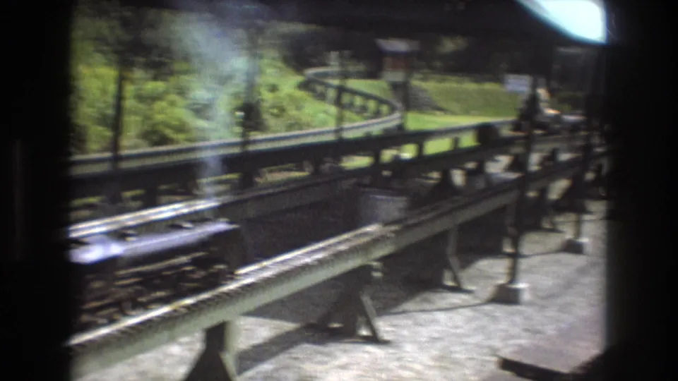 A small steam locomotive appears on curving miniature railway tracks in an outdoor 1975 Baxter, Maine home-movie view, with grass and trees beyond the rails.
