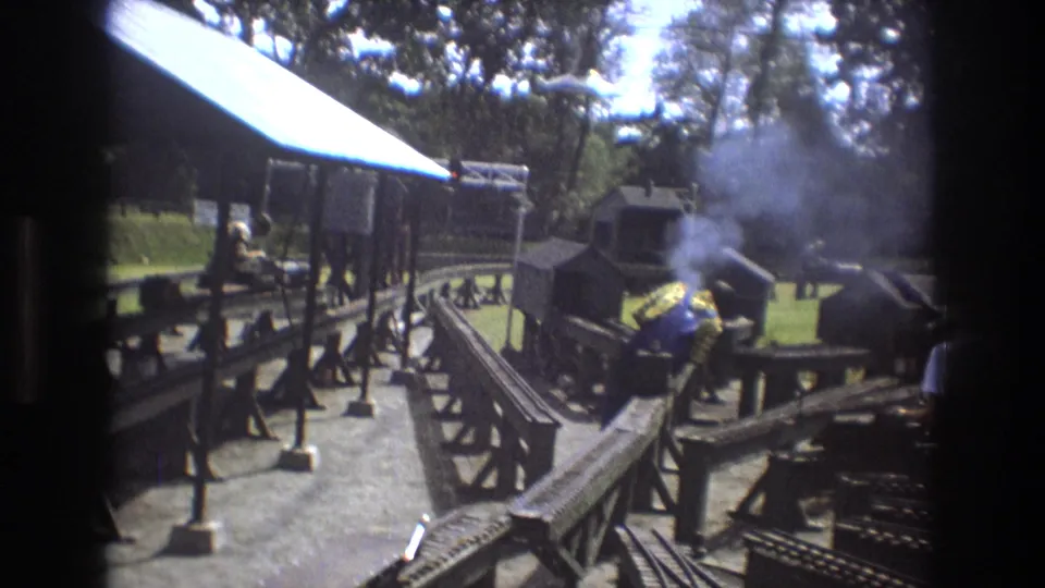 A small steam locomotive gives off smoke near a shaded structure and miniature track junction in the 1975 Baxter, Maine home-movie footage.
