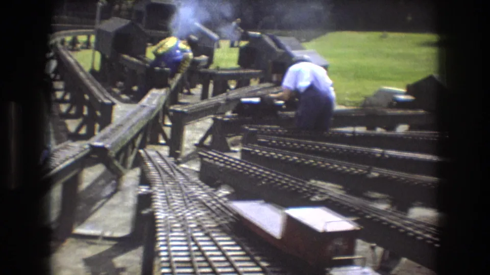 A man in a white shirt bends over a section of miniature railway track near a smoking small locomotive in this 1975 Baxter, Maine outdoor home-movie frame.