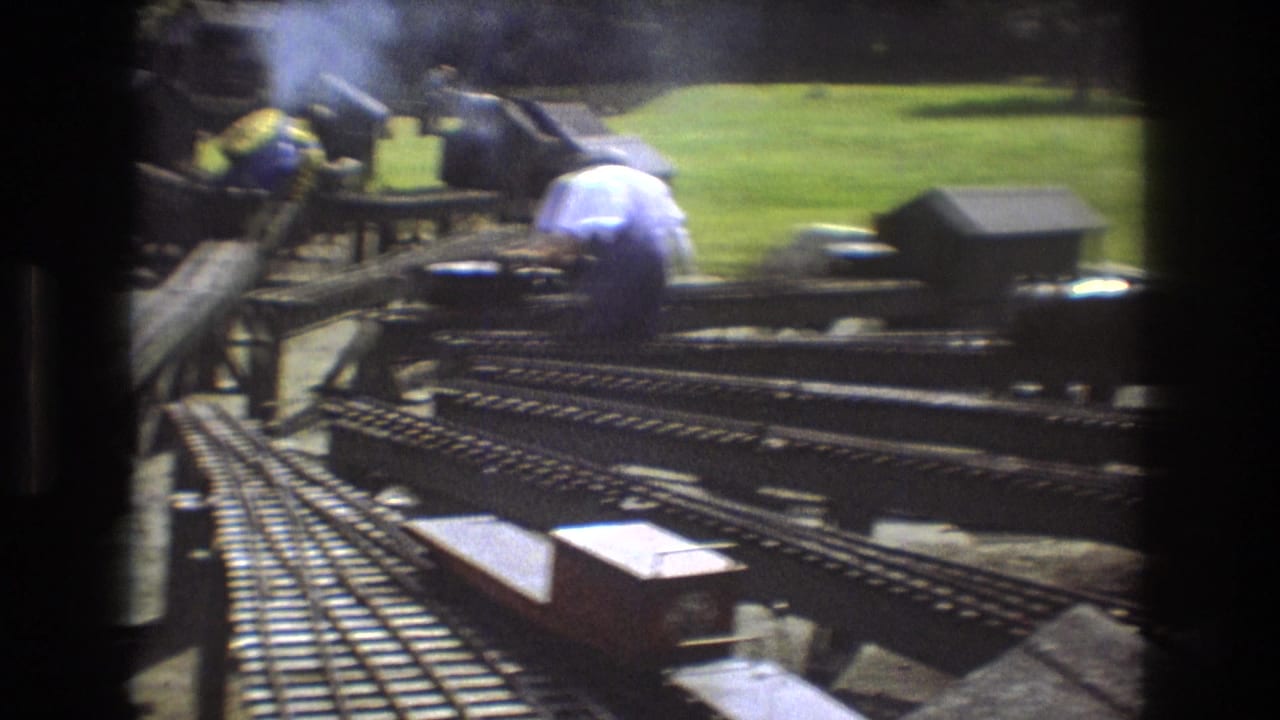1975 Baxter, Maine 8mm frame showing adults working around a complex outdoor miniature railroad layout with tracks, small structures, and grassy ground.