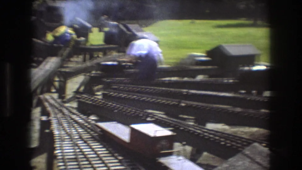 1975 Baxter, Maine 8mm frame showing adults working around a complex outdoor miniature railroad layout with tracks, small structures, and grassy ground.