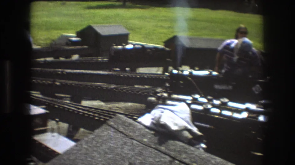 Outdoor 8mm view of a black miniature steam locomotive and track system with an adult standing near model structures on grass.