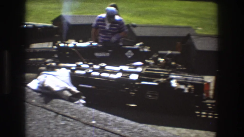 1975 home-movie frame of an adult in a striped shirt standing near a black miniature steam train, with tracks, equipment, and a white cloth in the foreground.