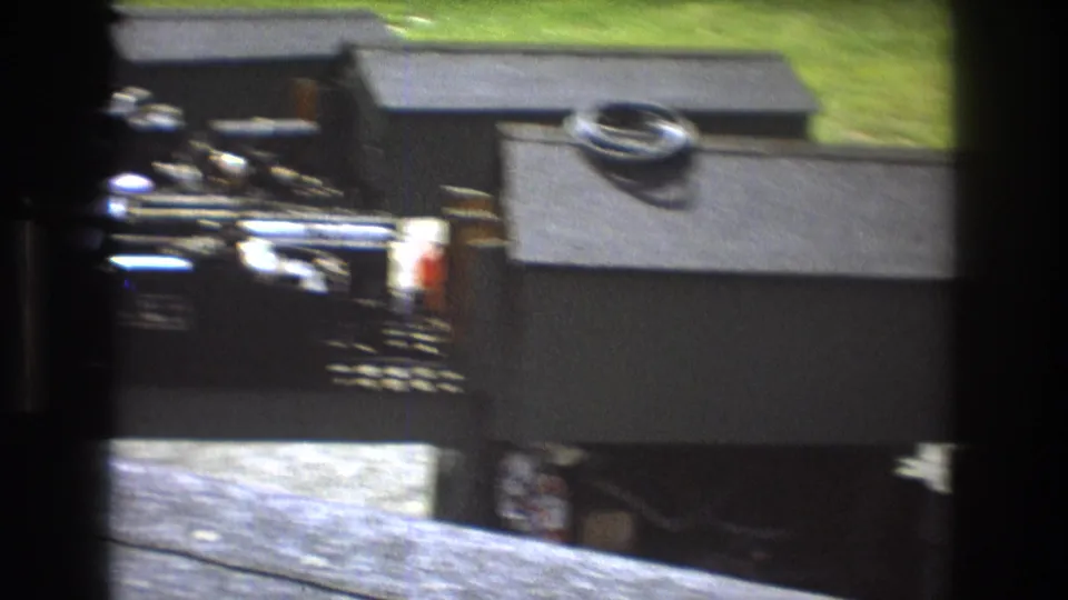 Soft 8mm frame showing a dark model railroad building, trackside equipment, a coiled object on the roof, and grassy background in the outdoor layout.