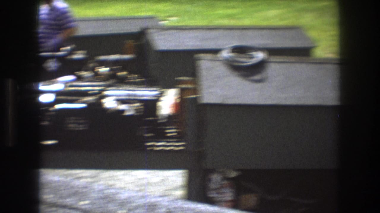 Baxter, Maine 1975 8mm frame with a black model locomotive partly obscured by miniature buildings and an adult standing near the outdoor display.