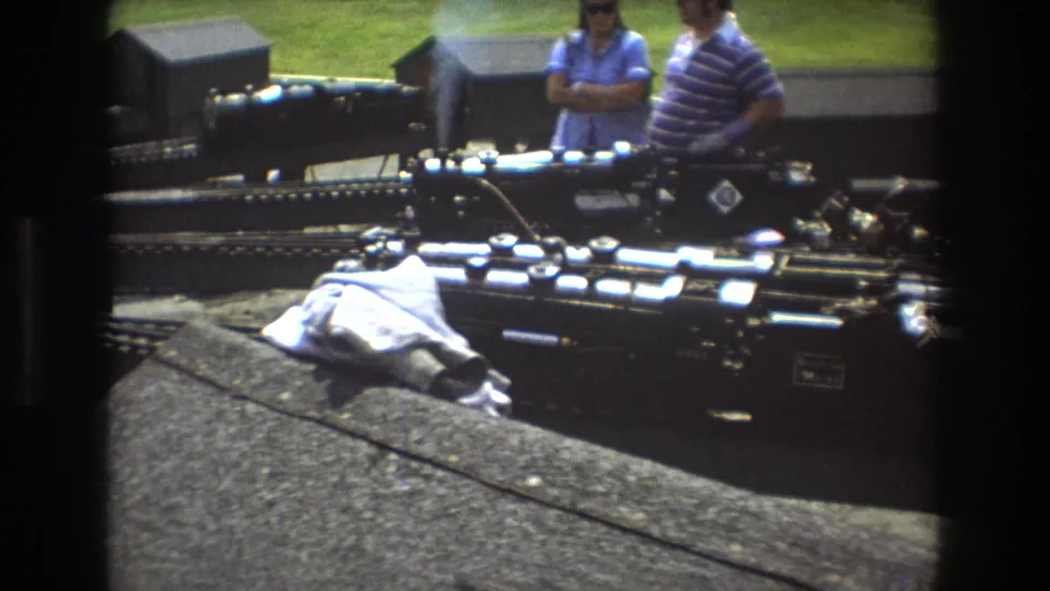Baxter, Maine 1975 8mm frame showing a black miniature steam locomotive on multiple tracks while two adults watch from behind the layout.