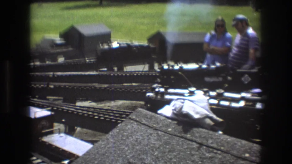 Baxter, Maine 1975 8mm frame showing smoke rising from a miniature train layout as two adults stand near the tracks at an outdoor exhibition.
