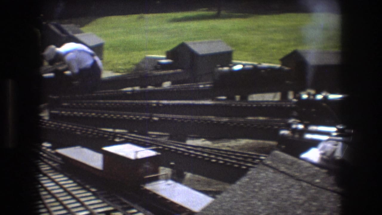 1975 Baxter, Maine 8mm frame showing an outdoor model railway with multiple tracks, small rolling stock, and an adult hobbyist leaning near the layout.