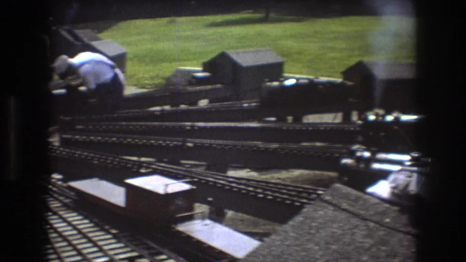 1975 Baxter, Maine 8mm frame showing an outdoor model railway with multiple tracks, small rolling stock, and an adult hobbyist leaning near the layout.