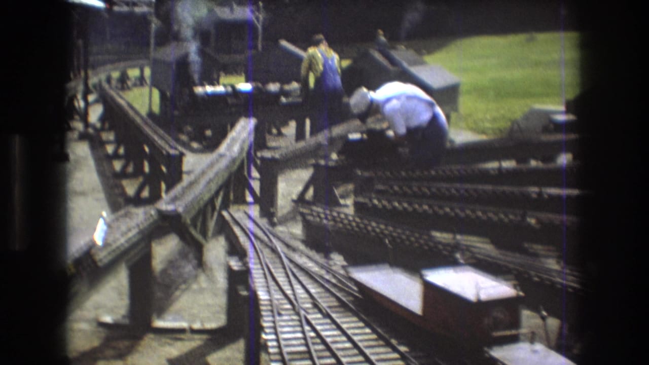 Smoking miniature steam locomotive on a model railway bridge with adults standing nearby.