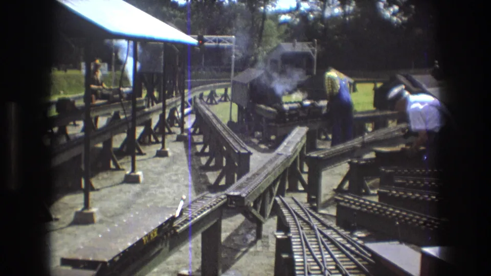 1975 Baxter, Maine 8mm frame showing a miniature steam locomotive producing smoke on an outdoor model railway, with bridgework, track switches, and adult hobbyists around the layout.