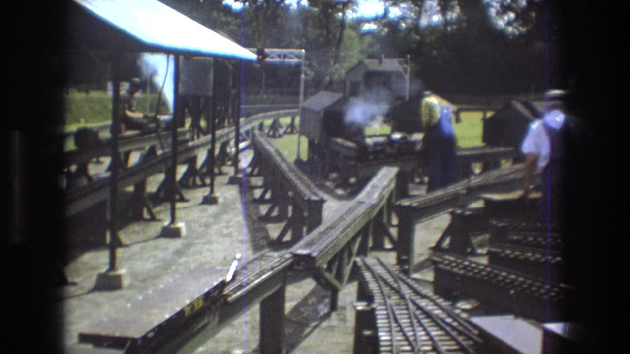 In Baxter, Maine in 1975, adult men work around a smoking miniature steam train station with raised tracks, platform supports, and trees in the background.