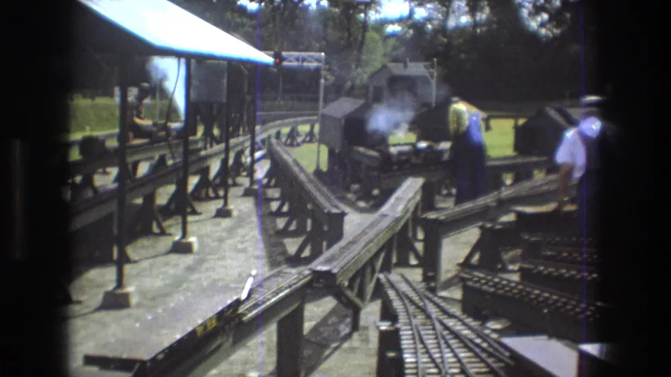 In Baxter, Maine in 1975, adult men work around a smoking miniature steam train station with raised tracks, platform supports, and trees in the background.