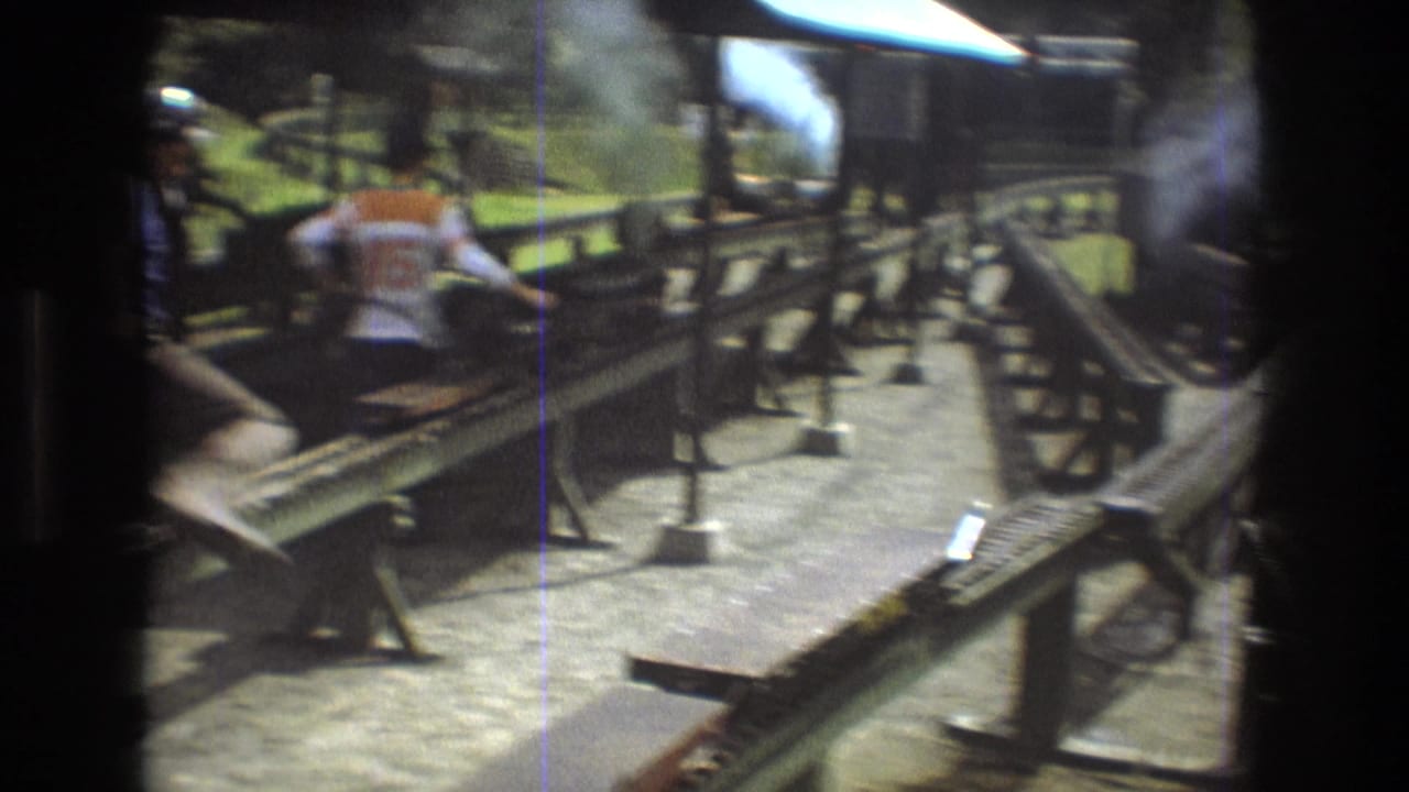 A man in a light shirt moves near raised miniature railway tracks under bright sun.