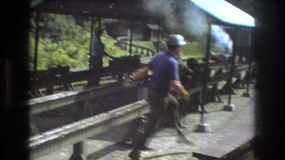In the Baxter, Maine 1975 footage, a man in a cap and blue shirt walks beside raised miniature train rails while smoke drifts from equipment near the platform.